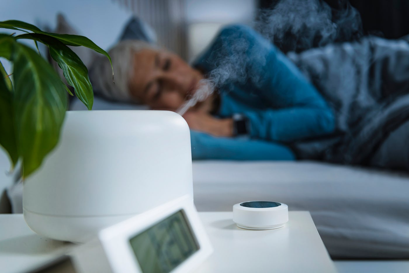 A cozy bedroom scene featuring a woman resting in bed while steam rises from a white humidifier. A plant and clock are on a bedside table.
