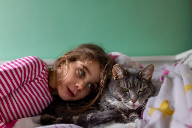 A girl with long brown hair lies beside a gray cat, both resting on a bed covered with colorful blan...