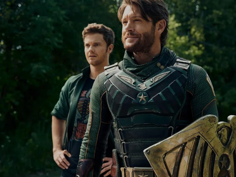 Two characters stand outdoors; one in a green, armored suit with a shield, and another casually dres...