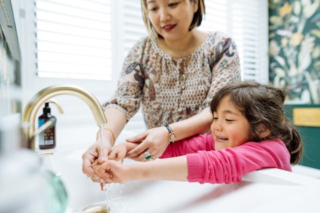 A woman and a young girl joyfully wash their hands at a sink. The girl, smiling, leans forward, whil...