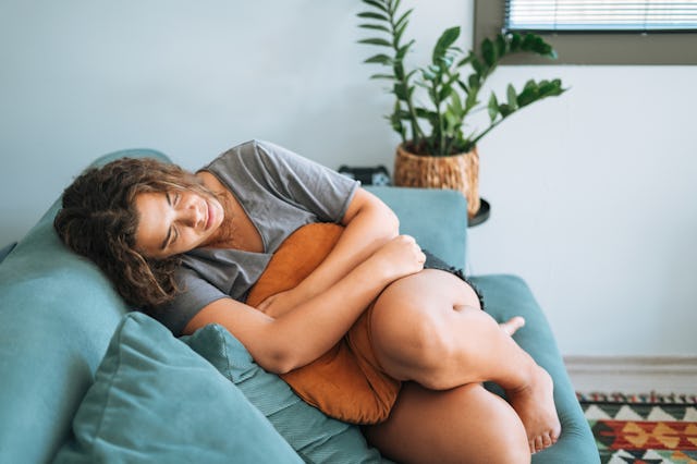 A woman sits on a teal couch, hugging a decorative pillow and resting her head peacefully. Green pla...
