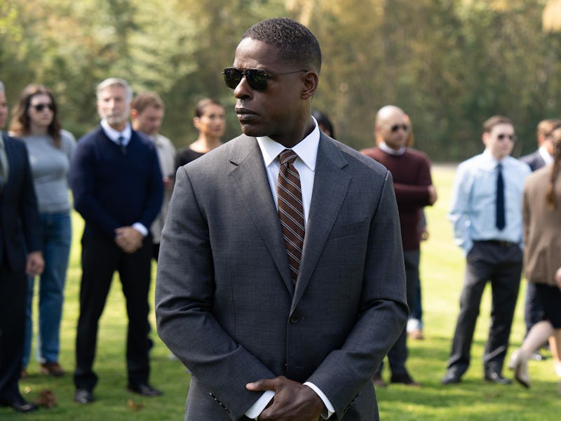 A man in a suit and sunglasses stands confidently in the foreground, surrounded by a group of people...