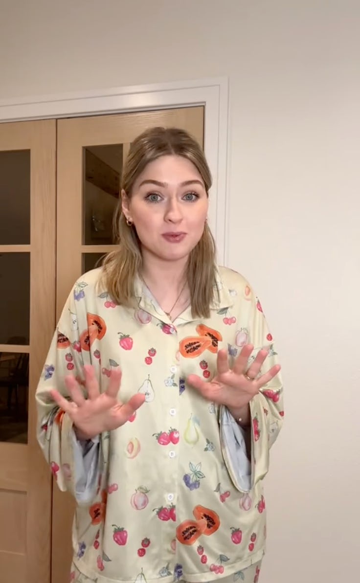 A young woman stands in front of a door, wearing colorful fruit-patterned pajamas. She gestures with her hands, expressing excitement or surprise.