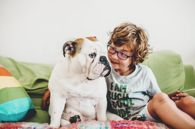 A child with curly hair and glasses sits on a couch, smiling at a bulldog beside him. Colorful pillo...