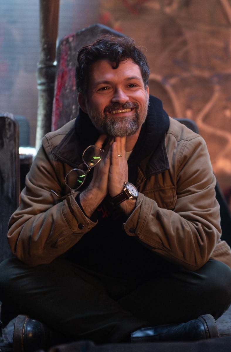 A man sitting cross-legged on the floor in a dimly lit, graffiti-covered room, smiling with his hand...
