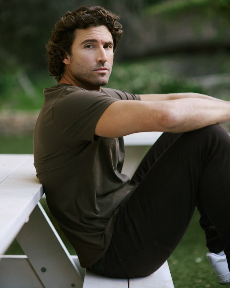 A man with curly hair sits on a picnic bench, gazing thoughtfully at the camera, dressed casually in…