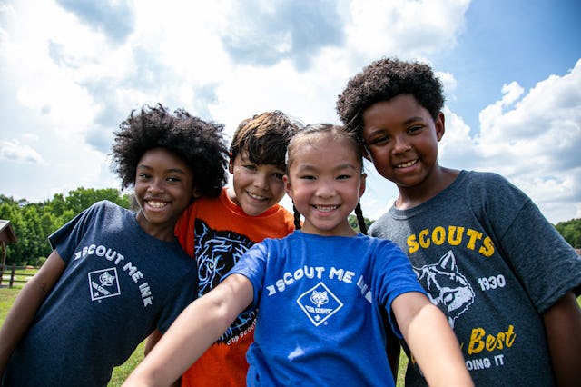 Four children smile at the camera outdoors, wearing colorful t-shirts. They pose playfully under a b...