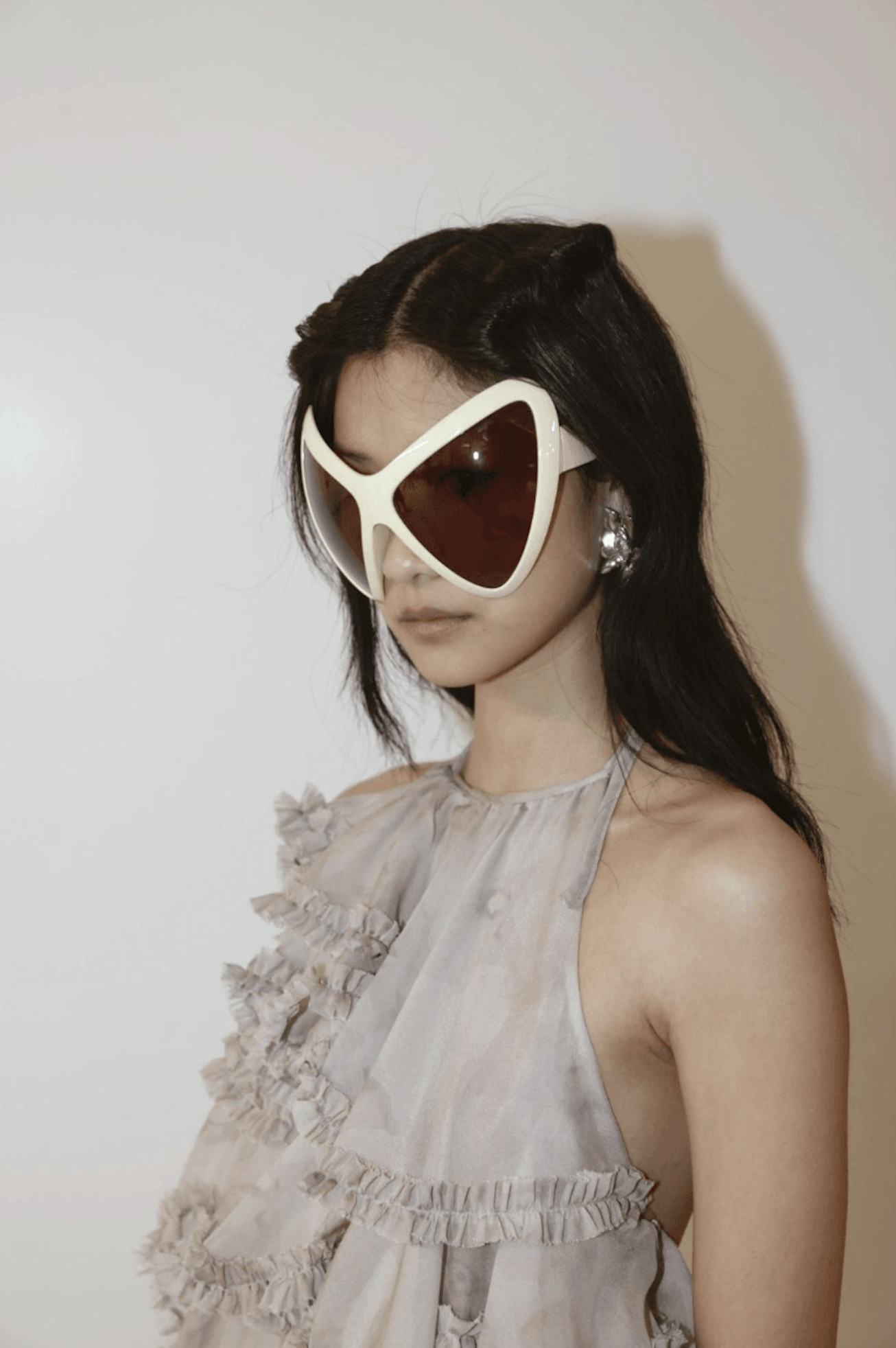 A model poses against a plain background, wearing oversized white and red sunglasses and a ruffled, ...