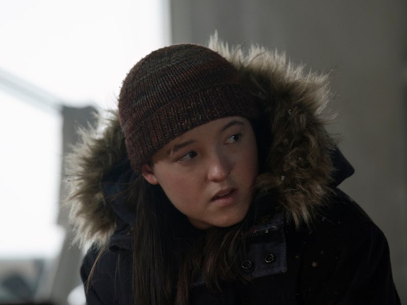 A young person wearing a dark winter coat with a fur-lined hood and a knit beanie looks thoughtfully...