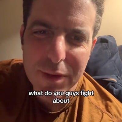 A close-up of a man with dark curly hair, sitting indoors with a soft light. He's looking directly at the camera, asking, "What do you guys fight about?"