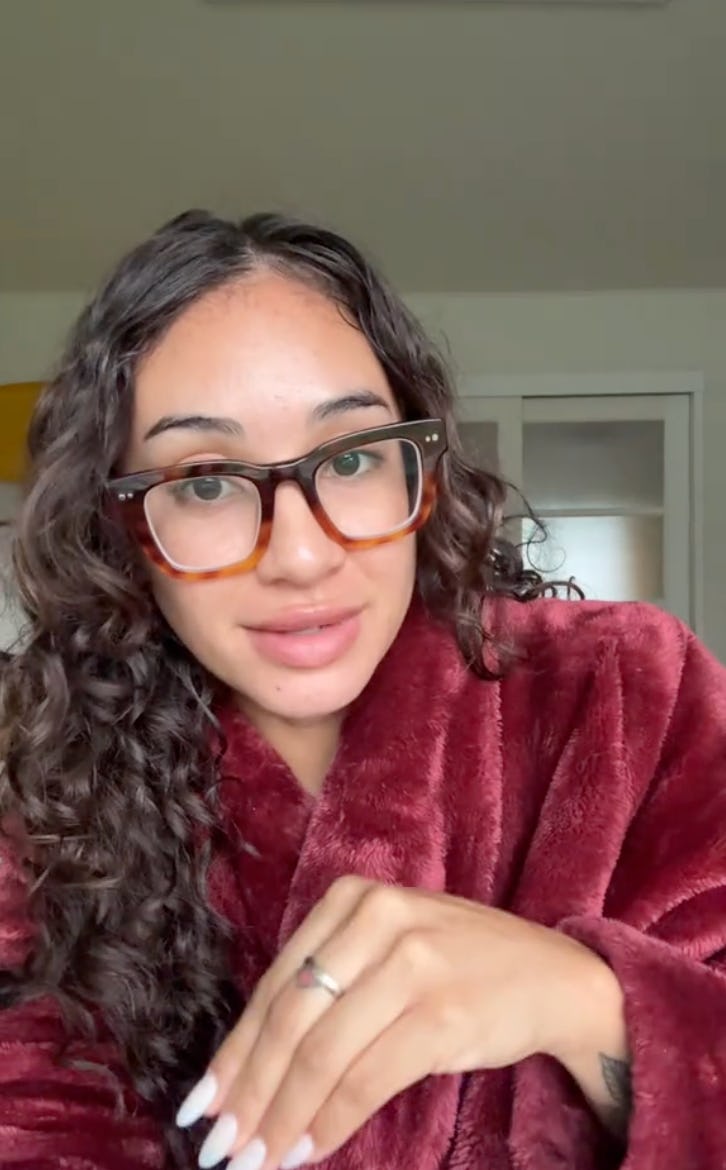 A young woman with curly hair wearing oversized glasses sits in a cozy, maroon robe. She is holding her hand up, smiling softly, in a bright room.
