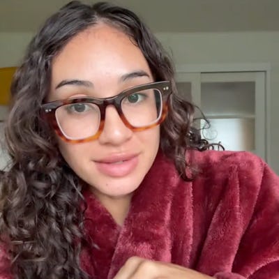 A young woman with curly hair wearing oversized glasses sits in a cozy, maroon robe. She is holding her hand up, smiling softly, in a bright room.