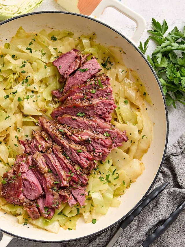 A close-up shot of sliced, seasoned corned beef atop sautéed cabbage, garnished with parsley in a wh...