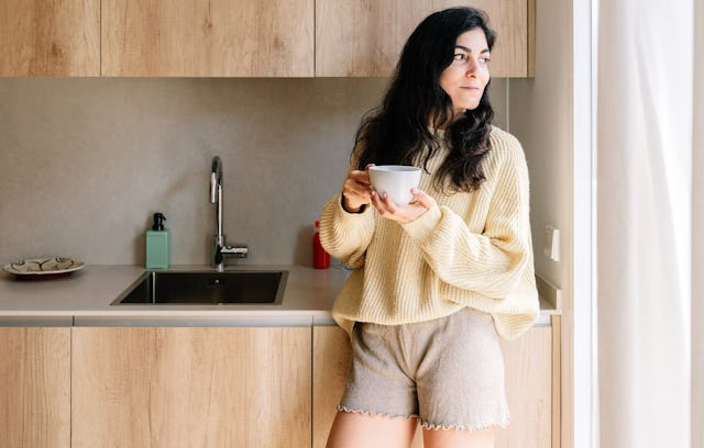 A woman in a cozy, oversized sweater and shorts stands in a modern kitchen, holding a cup, gazing th...