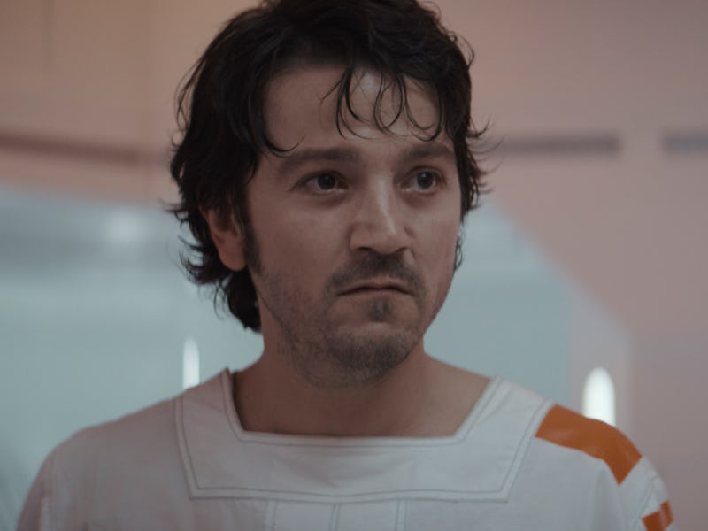 A man with dark hair and a serious expression wears a white space suit with orange accents, standing...