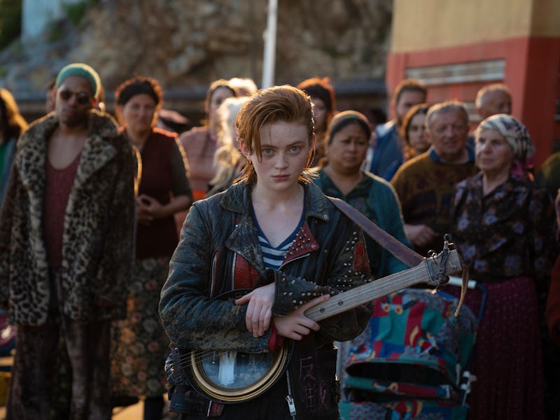 A young woman with tousled hair and a leather jacket stands in front of a crowd, holding a banjo-lik...