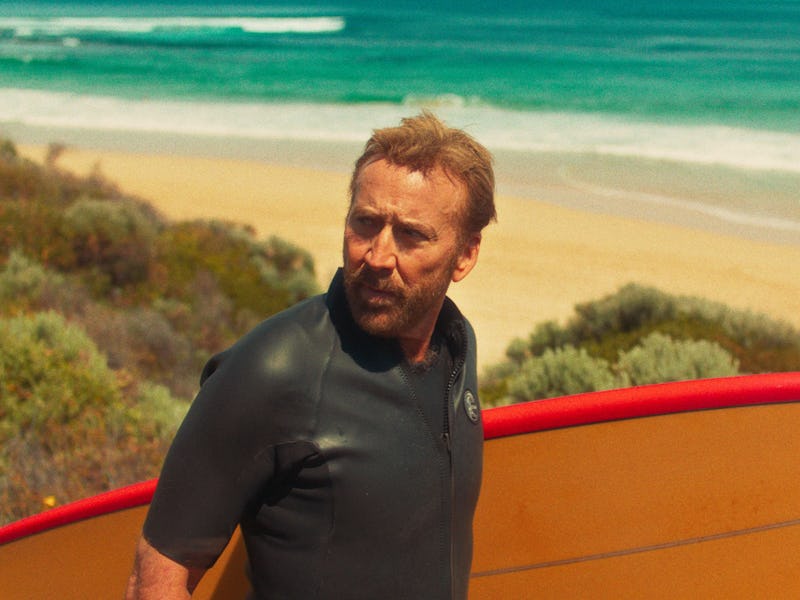 A man in a black wetsuit stands on a sandy beach, holding a red surfboard. He looks out toward the o...