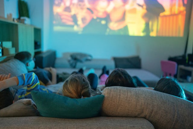 A group of four people sitting on a couch watching a movie projected on a wall, with soft lighting a...