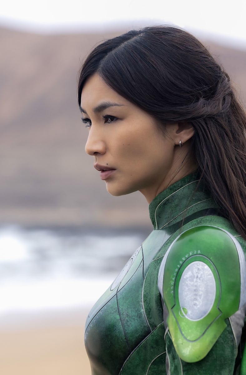 A woman in a green superhero costume stands profile against a beach backdrop, with waves and mountai...