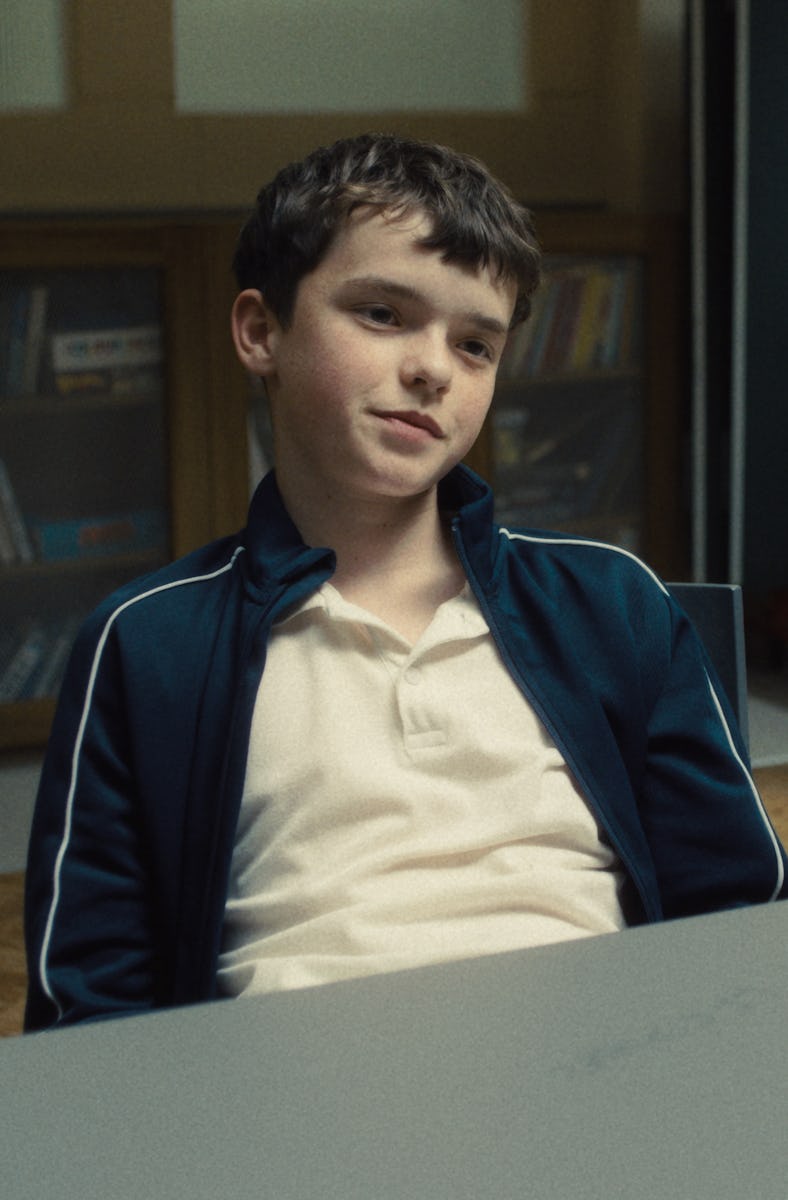 A young boy in a tracksuit sits at a table, looking relaxed. He is in a room with shelves of books a...