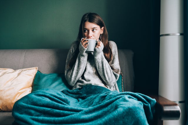 A young woman sits on a couch wrapped in a cozy teal blanket, holding a mug and looking thoughtfully...