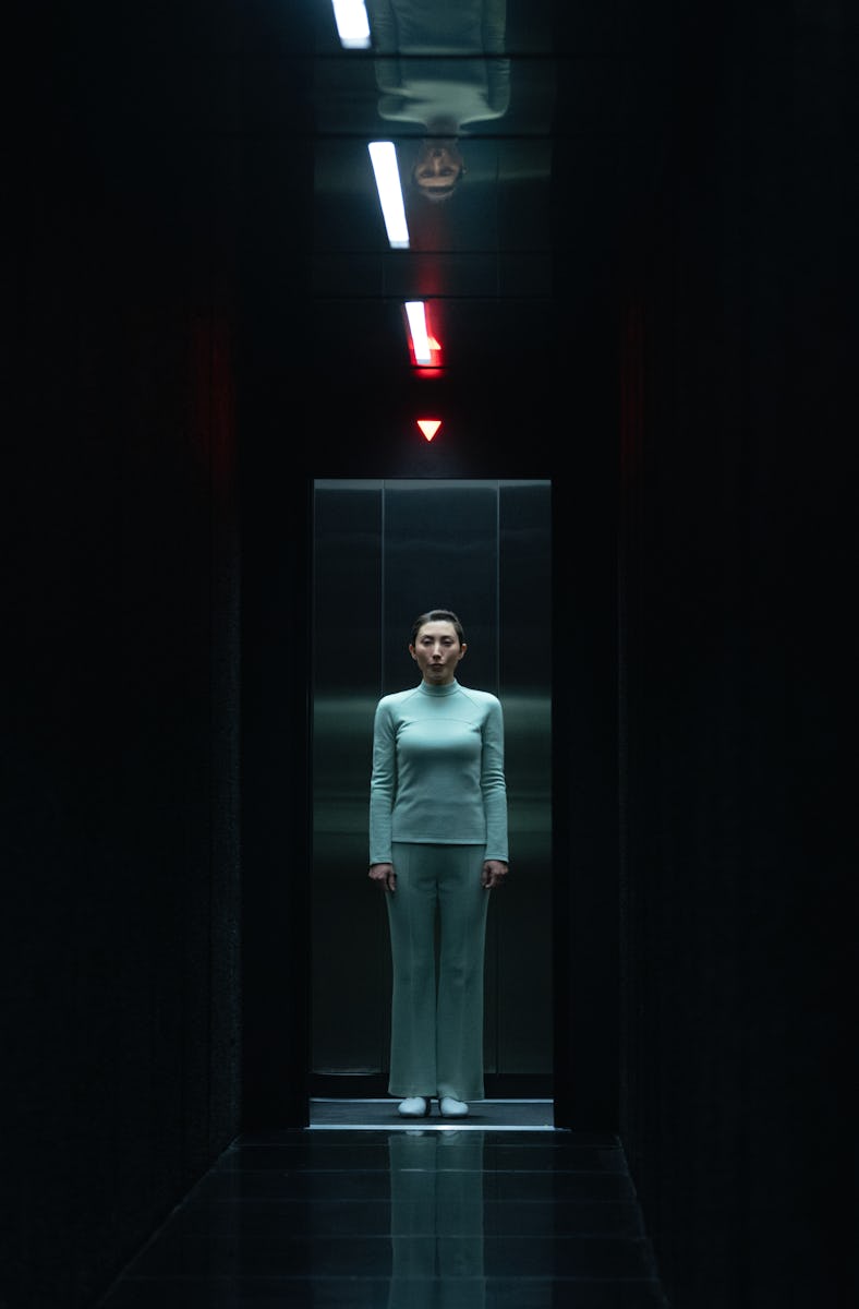 A dimly lit corridor features a woman in a pale outfit standing alone near an elevator, illuminated ...