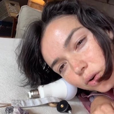 A woman with dark, tousled hair lies next to a light bulb and various objects, including crystals and a colorful parrot figure, on a textured surface.