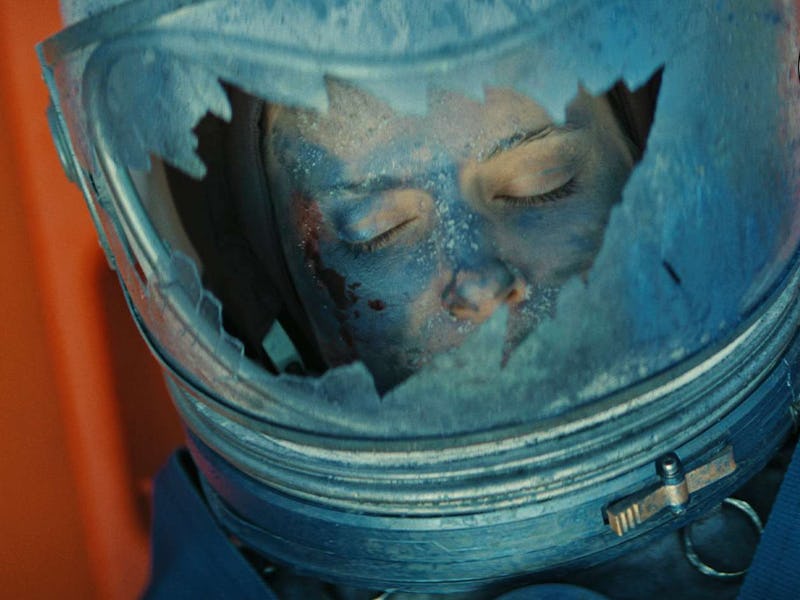 A close-up of a space helmet with a damaged visor, revealing a person's face with closed eyes, surro...