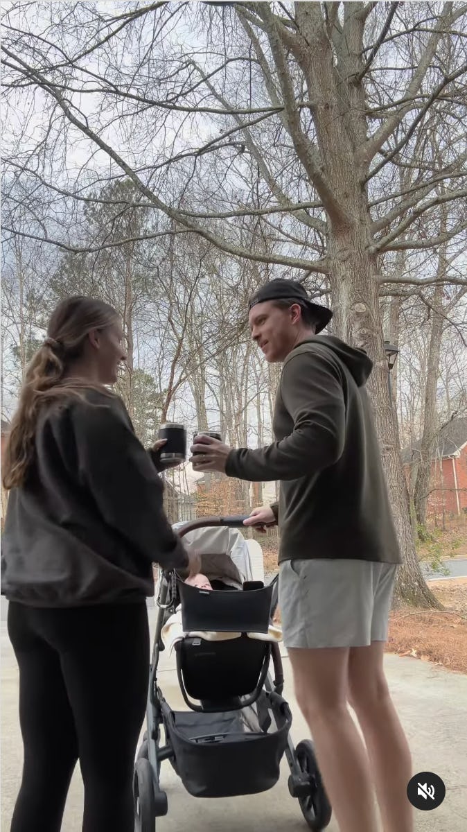 A woman and a man chat and laugh outside, each holding a drink. A stroller is nearby, with trees in the background, indicating a casual day out.