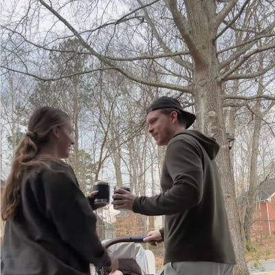 A woman and a man chat and laugh outside, each holding a drink. A stroller is nearby, with trees in the background, indicating a casual day out.
