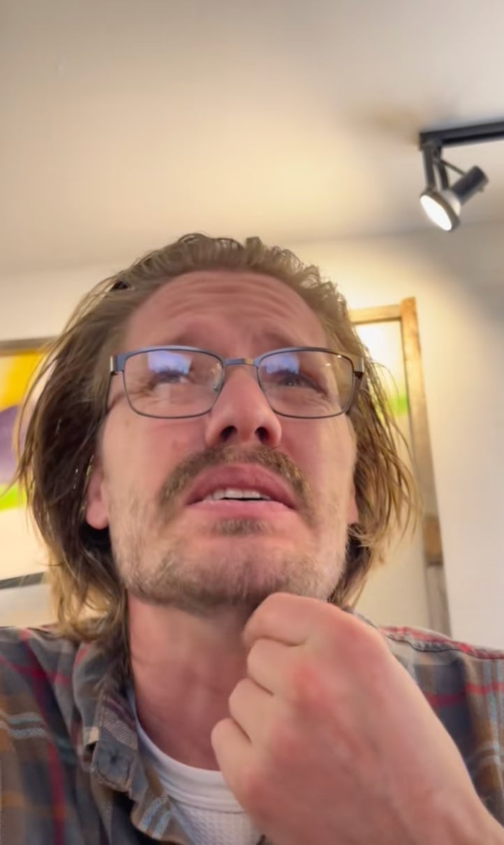 A man with long hair and glasses is seen from a low angle, looking up thoughtfully while resting his chin on his hand, in a cozy indoor setting.