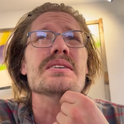 A man with long hair and glasses is seen from a low angle, looking up thoughtfully while resting his chin on his hand, in a cozy indoor setting.