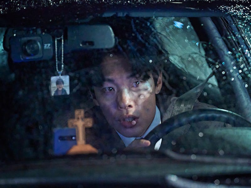 A young man looks intently through a rain-smeared windshield while driving at night. A small cross h...