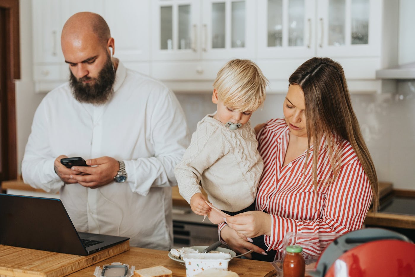 A family in a modern kitchen: a bearded man checks his phone, while a woman in a striped shirt holds a toddler, who examines a small item.