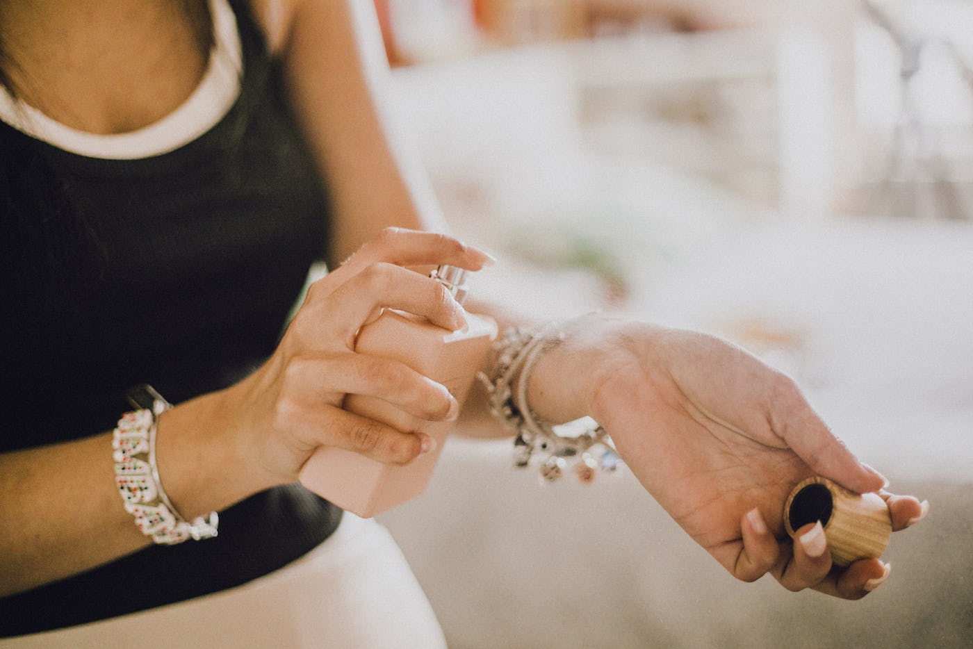 A close-up of a person applying perfume from a pink bottle onto their wrist, adorned with a charm bracelet, in a soft, blurred background.