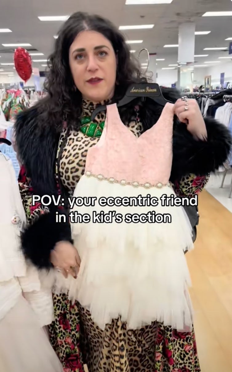 A woman in a stylish outfit, holding a pink and white dress from the kids' section, smiles while standing in a retail environment.