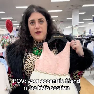 A woman in a stylish outfit, holding a pink and white dress from the kids' section, smiles while standing in a retail environment.