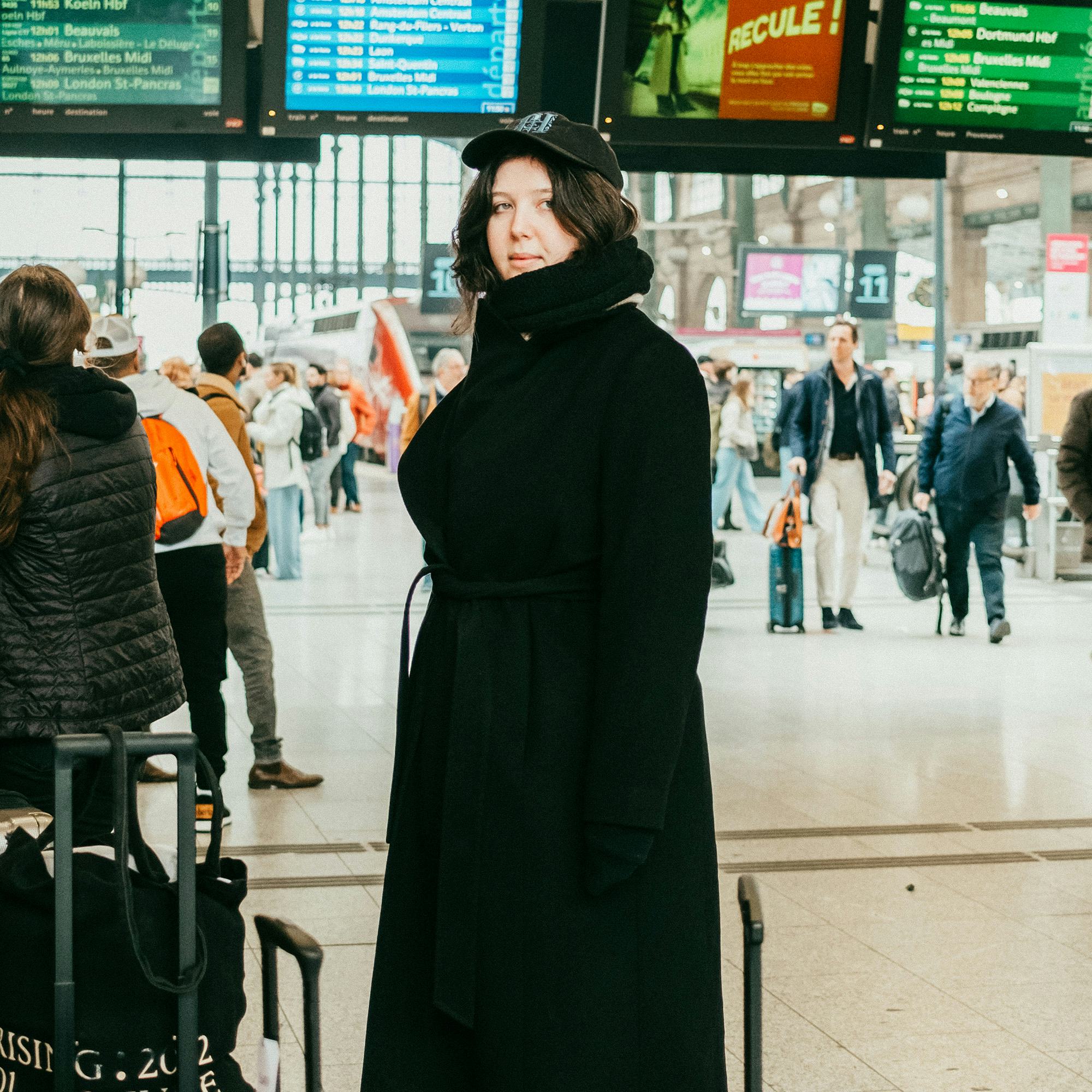 A young woman in a black coat and cap stands at a busy train station, surrounded by travelers and di...