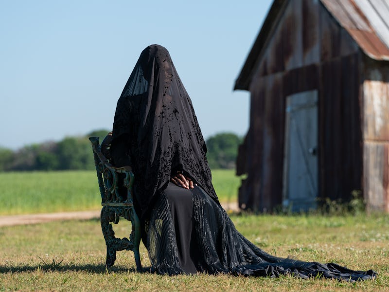 A figure in a long, flowing black garment and veil sits on a rustic chair in a grassy field, near a ...