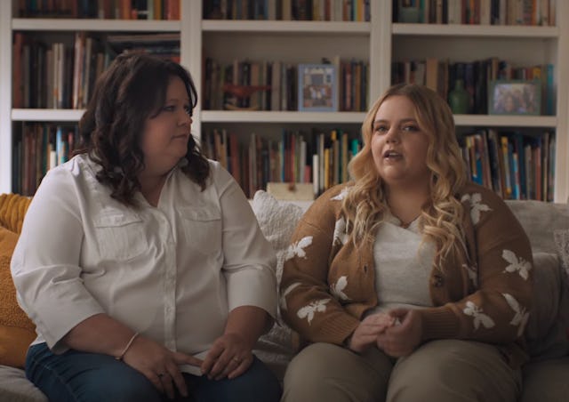 Two women sit on a couch in a cozy living room, surrounded by bookshelves. One woman is speaking whi...
