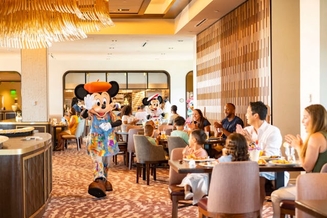 A lively restaurant scene features Mickey and Minnie Mouse in colorful attire, engaging with familie...