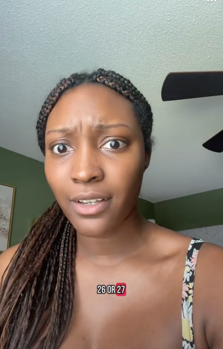 A young woman with braided hair looks surprised and questioning. She is indoors with a green wall backdrop, wearing a sleeveless floral top.