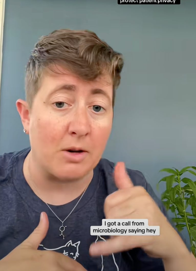 A person with short hair is speaking, gesturing as they recount receiving a call from microbiology. They wear a casual shirt, and a plant is in the background.