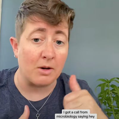 A person with short hair is speaking, gesturing as they recount receiving a call from microbiology. They wear a casual shirt, and a plant is in the background.