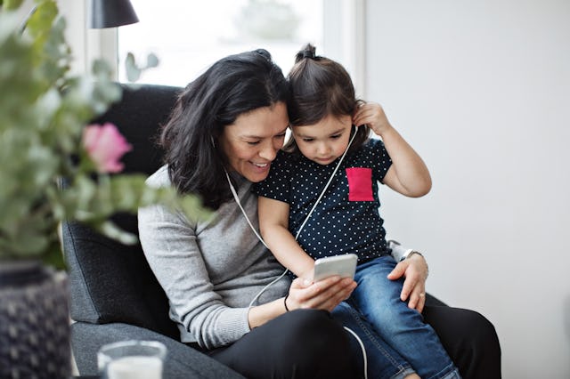 A smiling woman and a young girl sit together on a couch, sharing earbuds and looking at a smartphon...