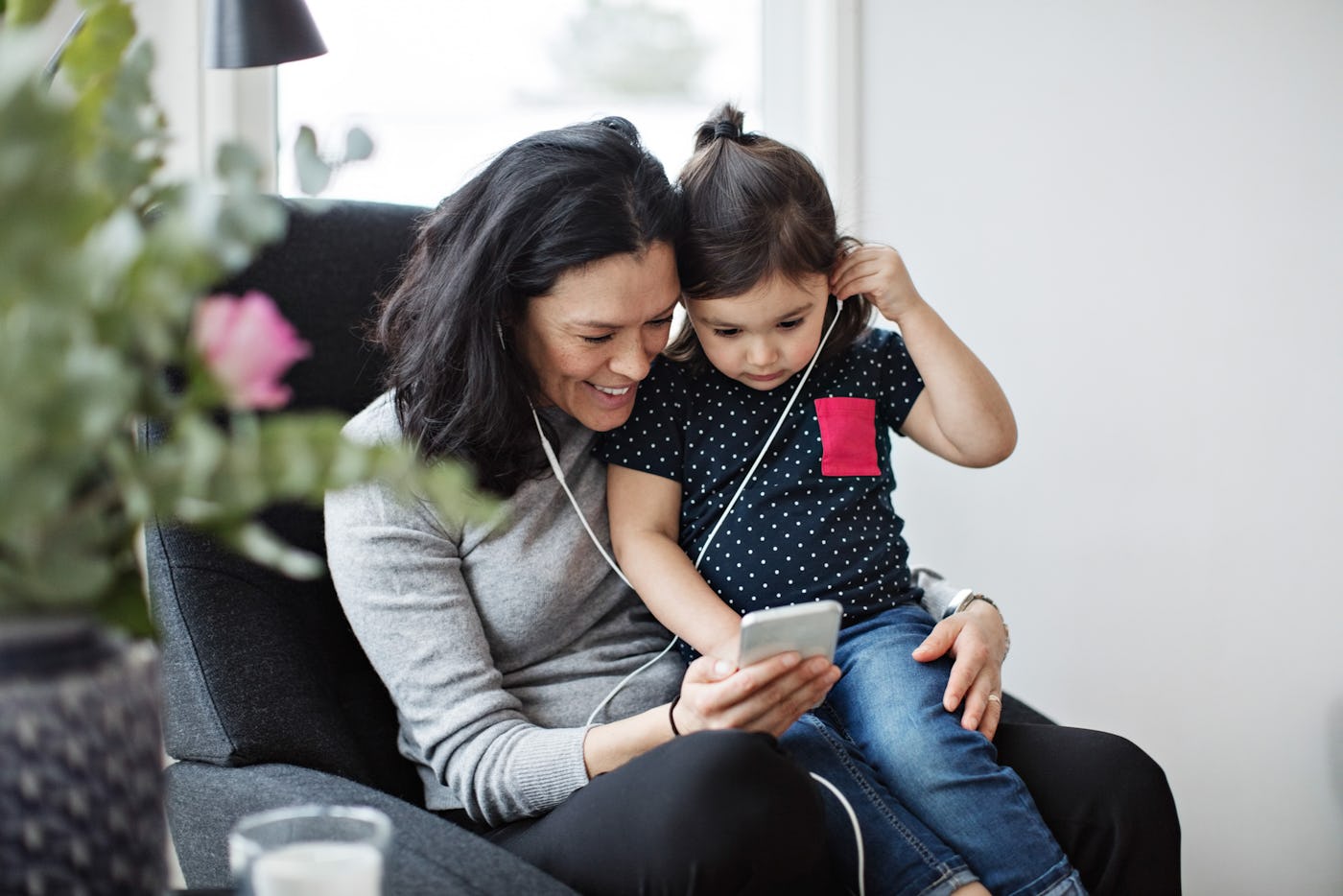A smiling woman and a young girl sit together on a couch, sharing earbuds and looking at a smartphone, surrounded by a cozy living room setting.