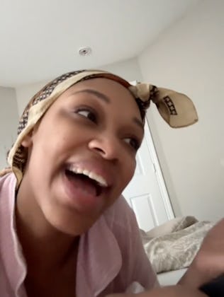 A woman with a headscarf smiles and talks animatedly while lounging in bed, surrounded by soft pillo...