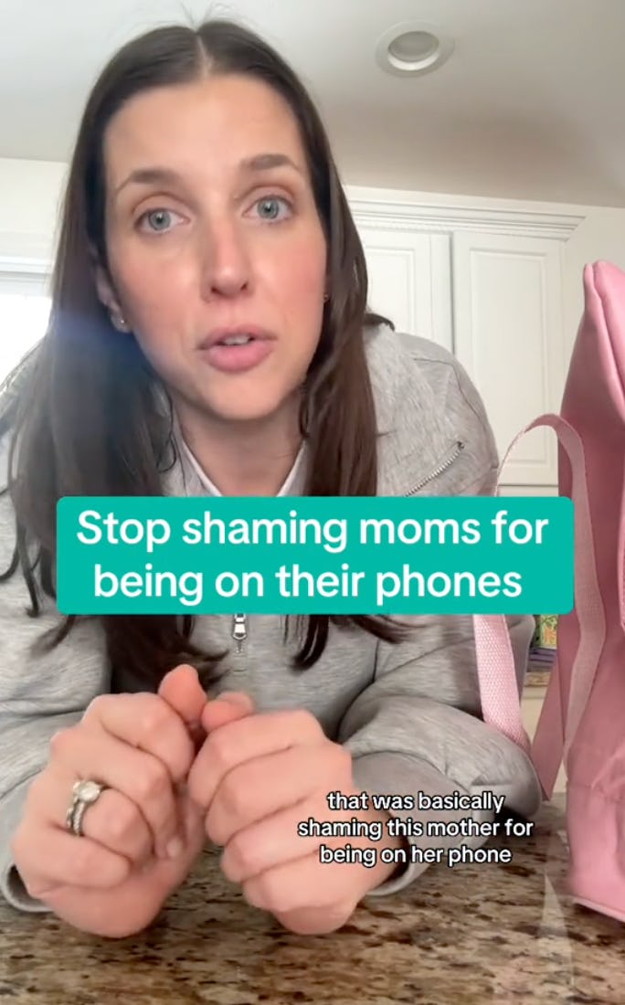 A woman with long brown hair sits at a table, looking directly at the camera. Text on the image reads: "Stop shaming moms for being on their phones."
