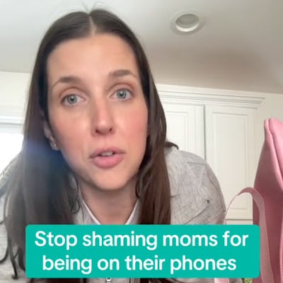 A woman with long brown hair sits at a table, looking directly at the camera. Text on the image reads: "Stop shaming moms for being on their phones."