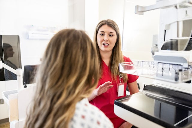 A healthcare professional in red scrubs speaks with a patient in a medical setting. The patient wear...